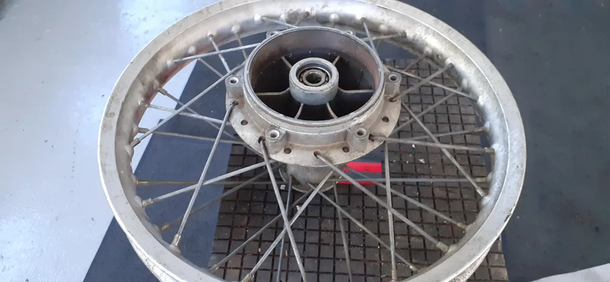 A close-up photo of a motorcycle wheel without a tire, showing the metal rim, spokes, and central hub assembly resting on a…