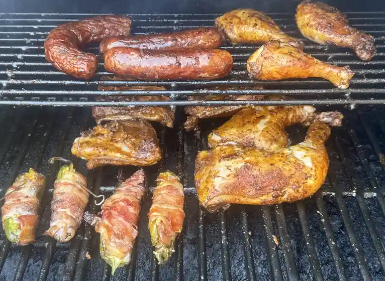 A close-up photo of a grill filled with various meats including sausages, chicken legs, ribs, whole chickens, and…