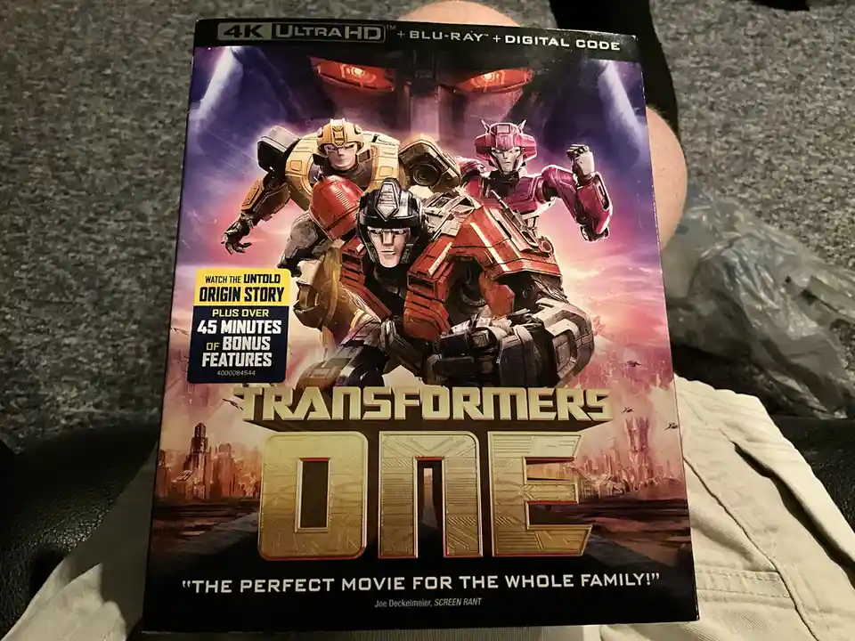 A person holds a 'Transformers One' 4K Ultra HD Blu-ray box set featuring three robot characters on the cover. The box…