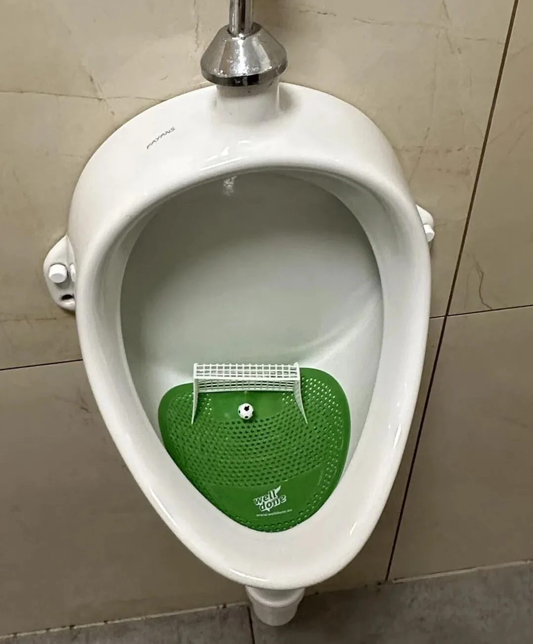 A white urinal mounted on tiled walls contains a green, perforated mat with a miniature white soccer goal and a tiny…