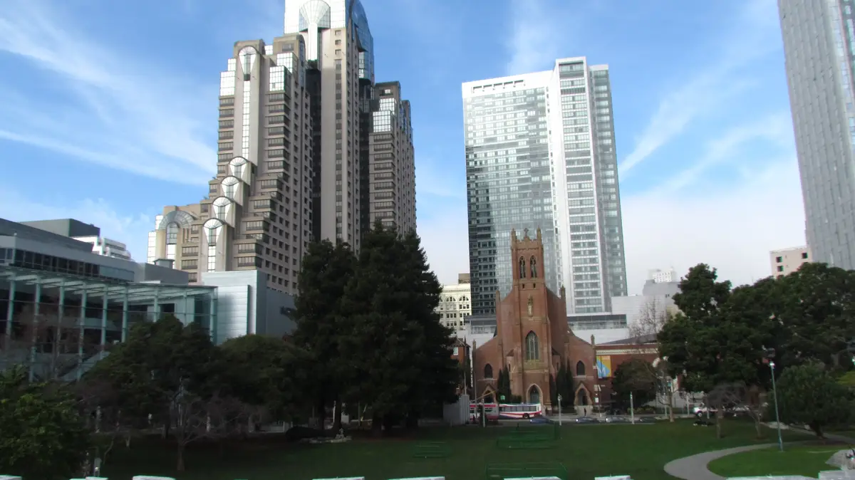 A city park scene with a small red brick church in the foreground surrounded by green grass and trees, with several tall…