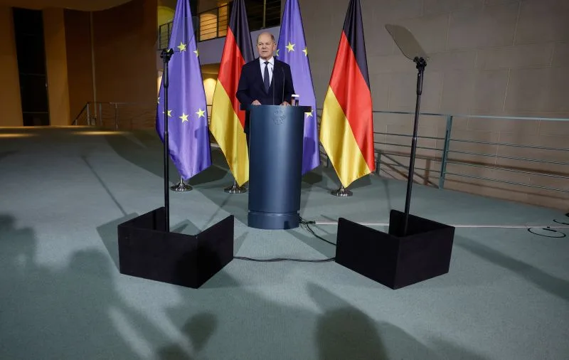 a man in a suit stands at a podium flanked by two european union flags and two german flags. The image is overlaid with the…