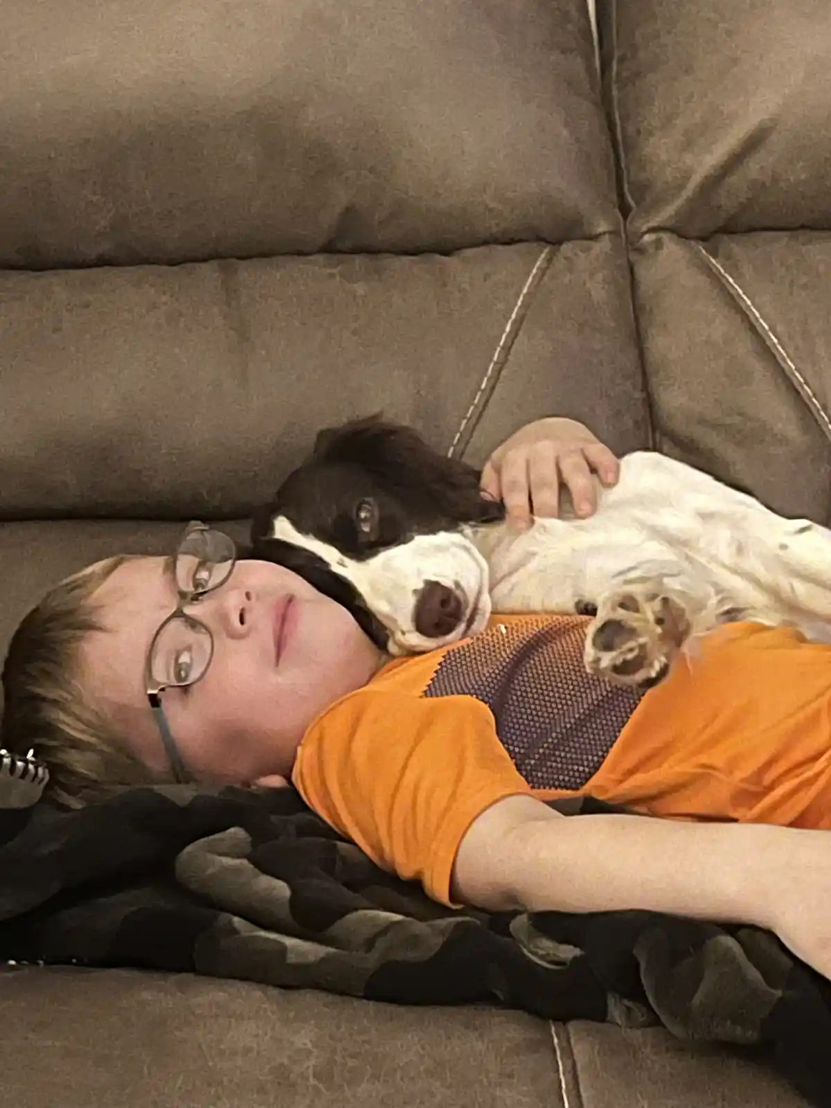 A young boy with glasses lies on a brown couch, wearing an orange shirt, with a black-and-white dog resting its head on his…