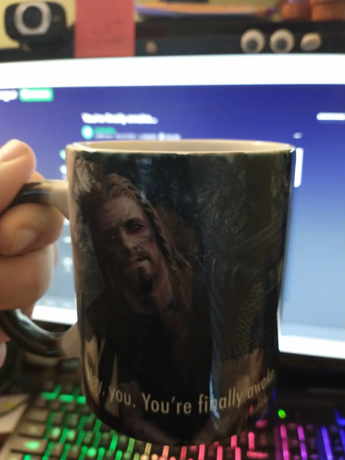 A hand holds a mug featuring a close-up image of a weary-looking man with long hair and a beard, overlaid with the text…