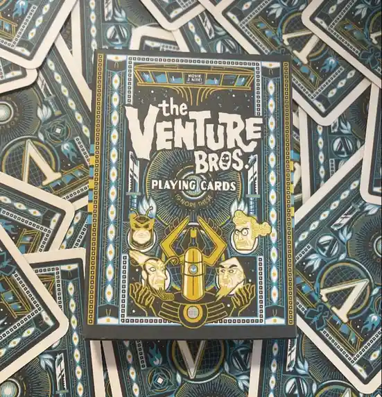 A box of 'The Venture Bros. Playing Cards' sits atop a scattered pile of the same cards, featuring stylized illustrations of…