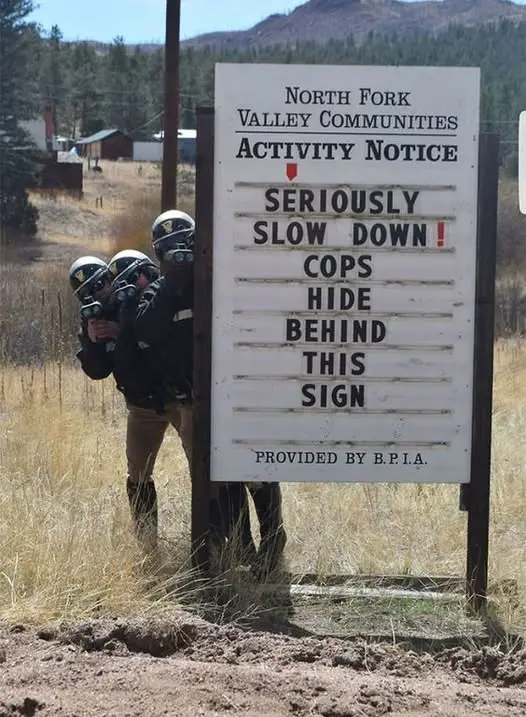 Three police officers in full gear are peeking out from behind a large white sign that reads 'NORTH FORK VALLEY COMMUNITIES…