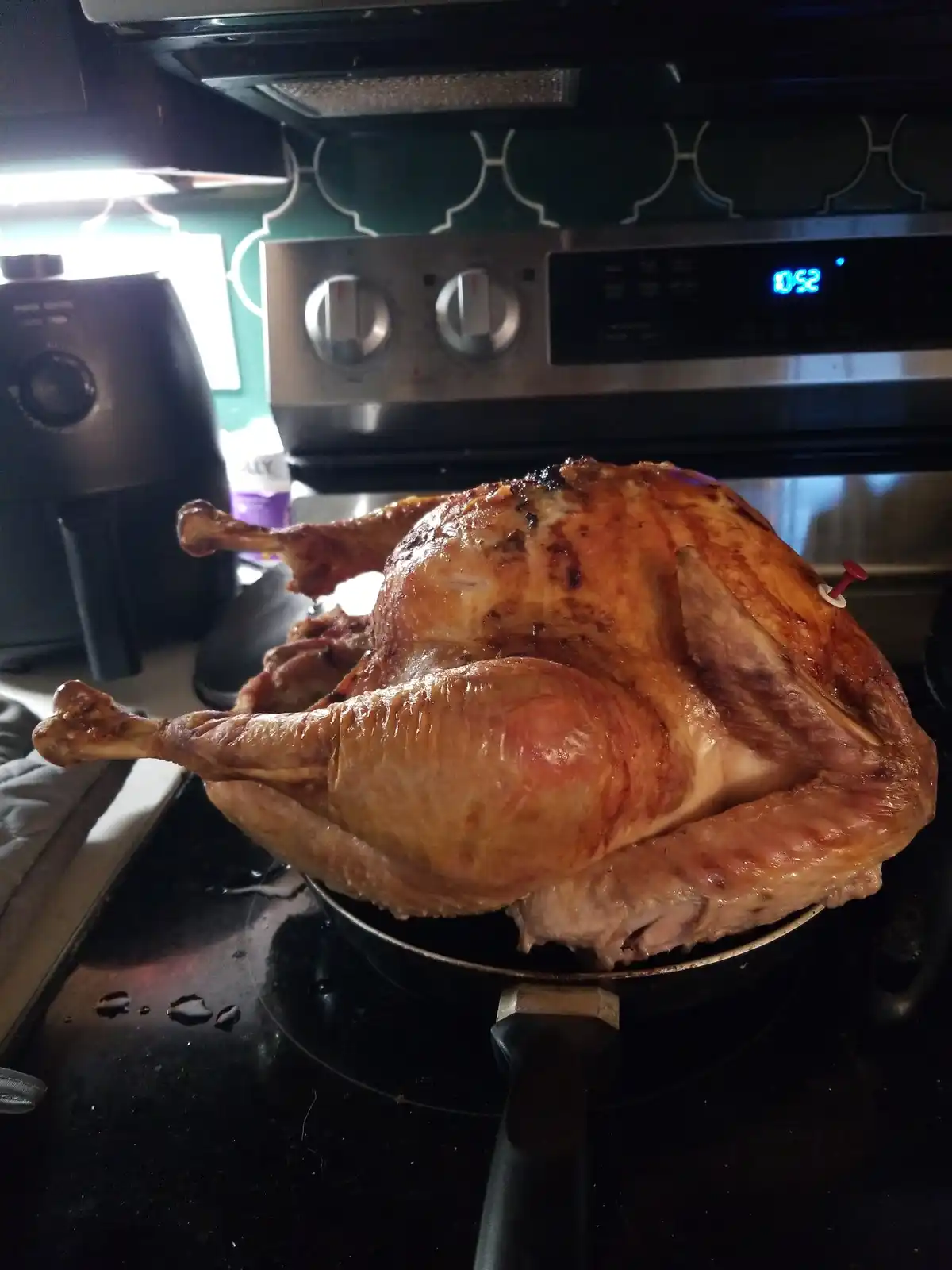 A roasted turkey sits in a pan on a stovetop, its skin golden-brown and crispy. The oven's digital display clearly shows…