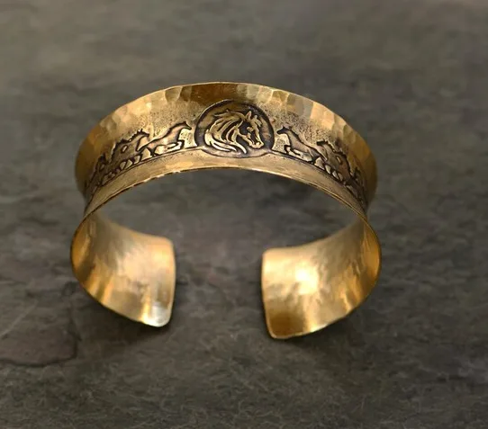 A golden, hammered metal cuff bracelet with an engraved horse head and mountain range design rests on a dark slate surface…