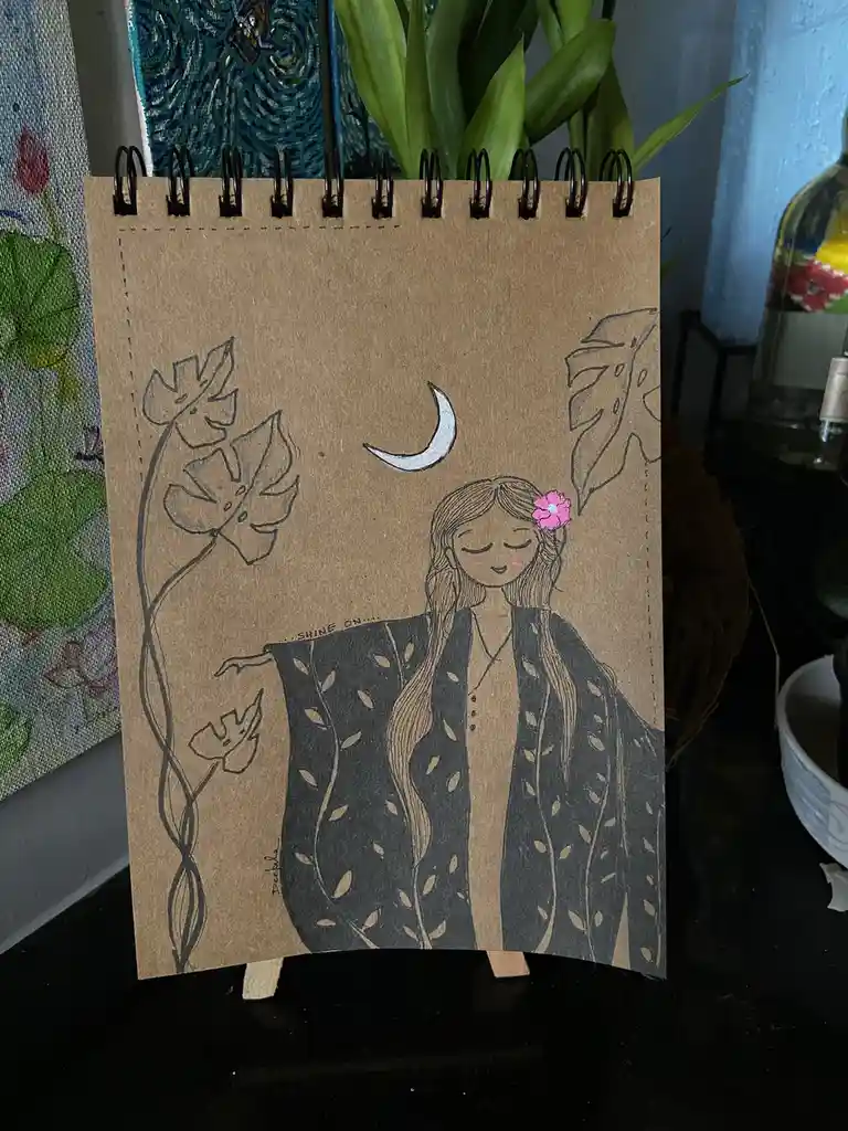 A hand-drawn illustration on brown kraft paper depicts a serene woman with long hair and a pink flower in her hair, wearing…