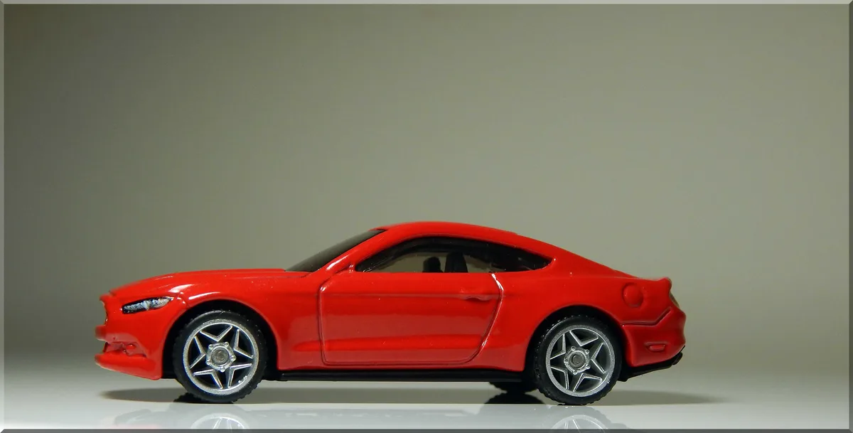 A red toy car, resembling a Ford Mustang, is shown in profile against a plain gray background. The car has silver…