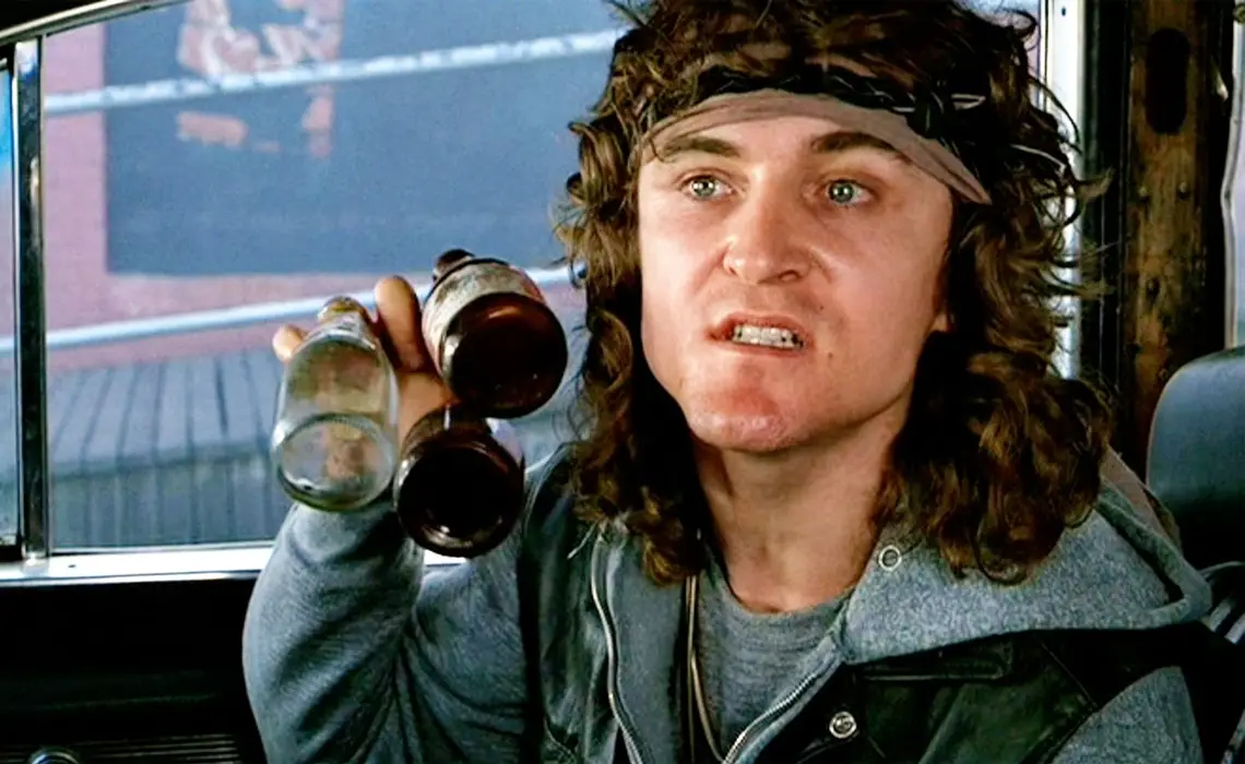 a man with long curly hair and a headband sits inside a vehicle, holding up three empty glass bottles, looking slightly…
