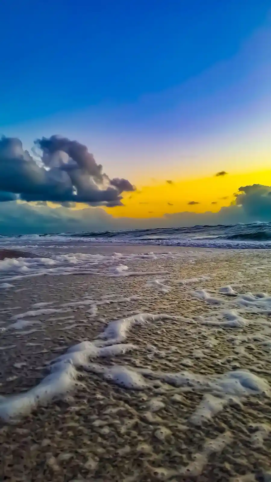A serene beach scene at sunset with waves washing over sand, featuring a dramatic sky with blue and yellow hues and clouds…