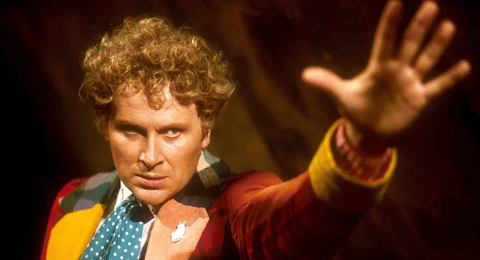 A man with curly hair, wearing a colorful jacket and a blue polka-dot tie, holds his hand out with a serious expression. The…