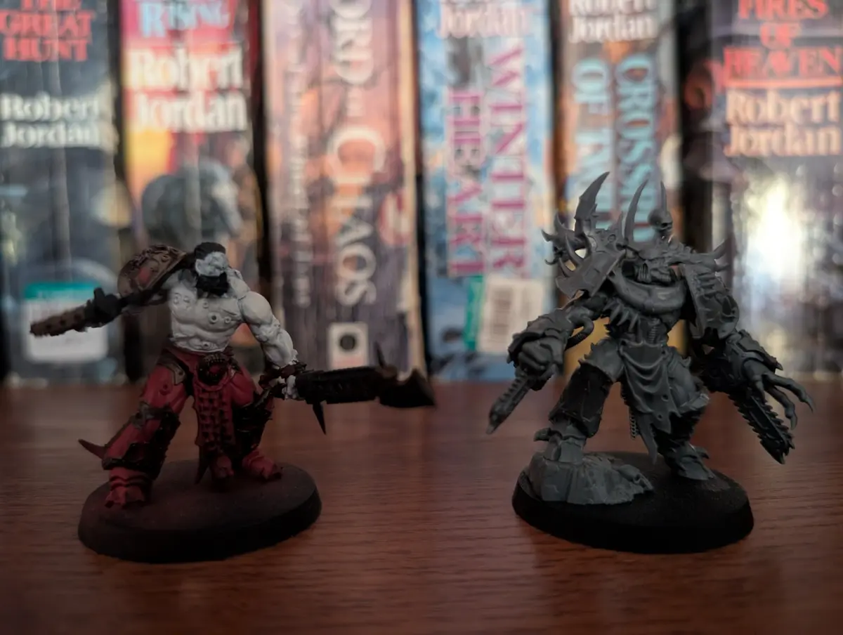 Two painted Warhammer 40k miniatures face each other on a wooden surface, one in red and grey armor holding a chainsword…