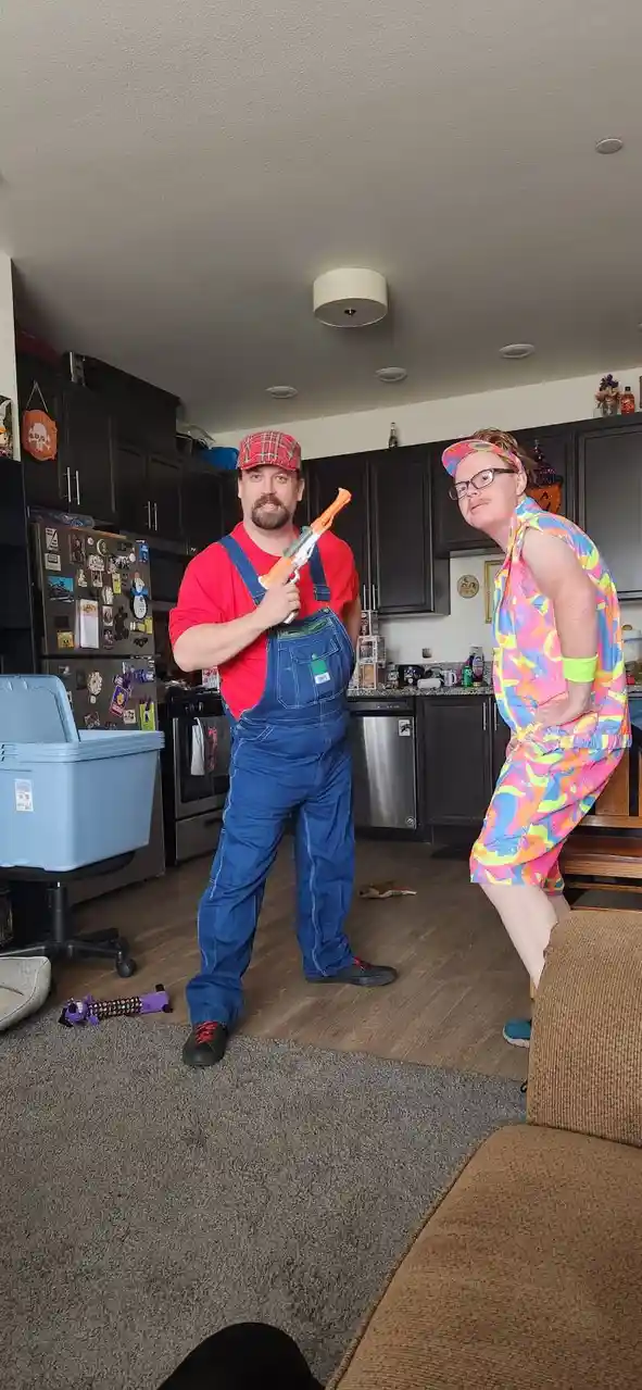 Two people pose in a kitchen/living room; one wears a red shirt and blue overalls holding an orange toy gun, the other wears…