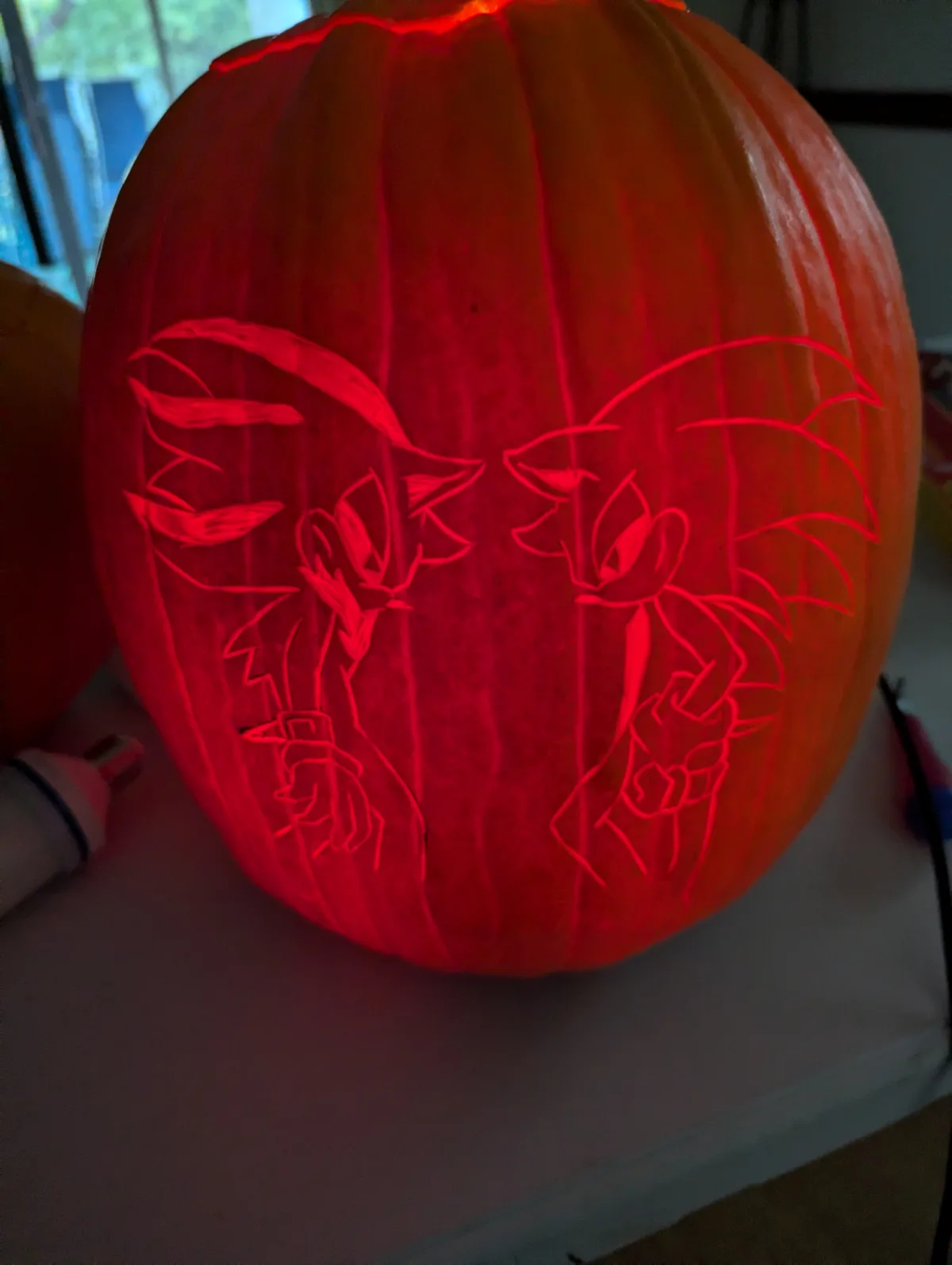 A carved pumpkin illuminated from within, featuring two stylized Sonic the Hedgehog characters facing each other with their…