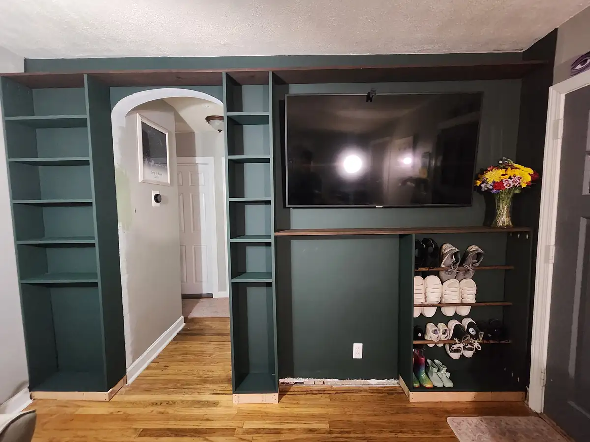 A photo of a newly built green wall unit with built-in shelves, a mounted TV, and a shoe rack. The TV screen reflects two…