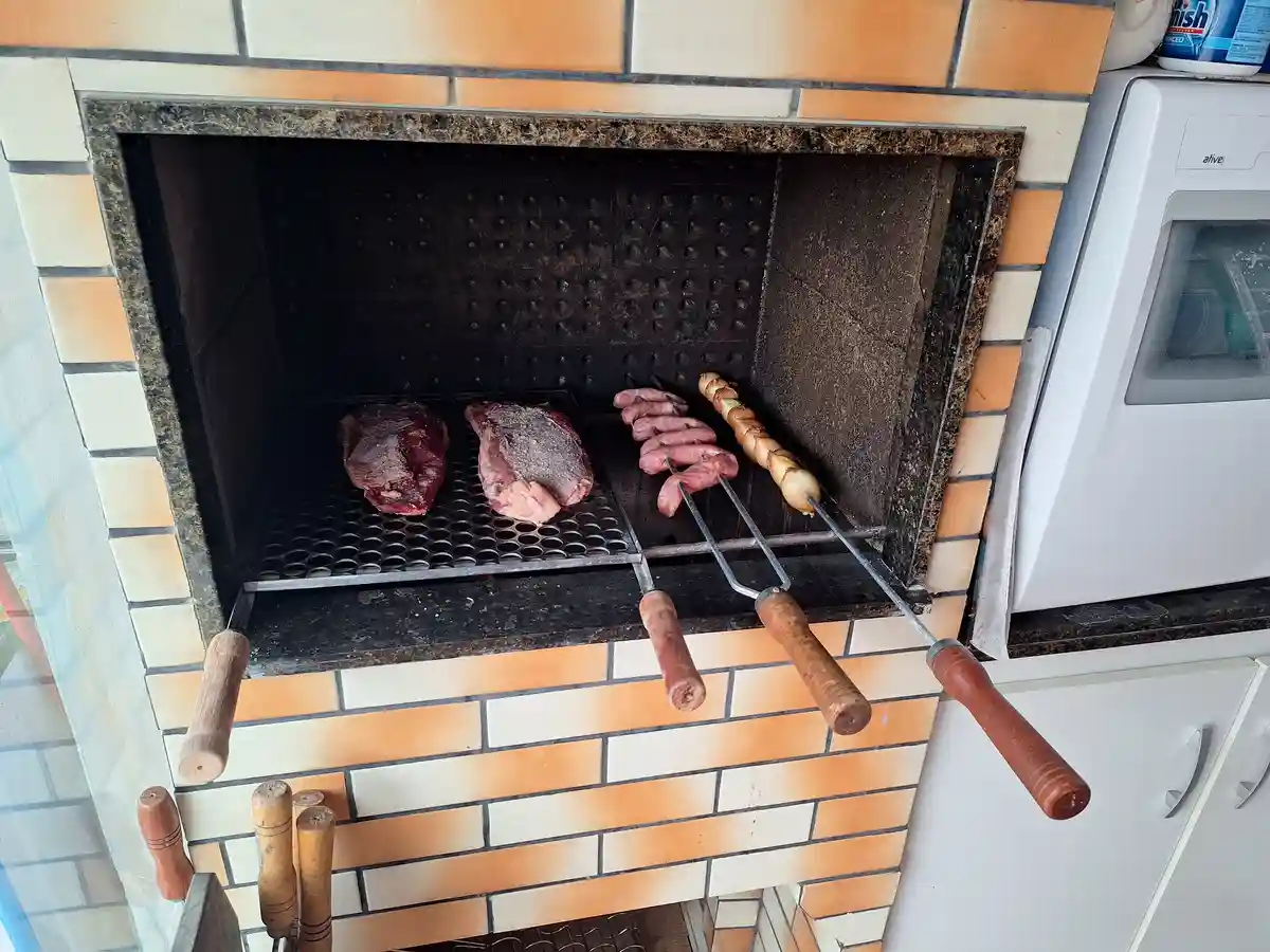 Raw meat, including steaks and sausages on skewers, is being cooked inside a brick fireplace with a metal grate. The…