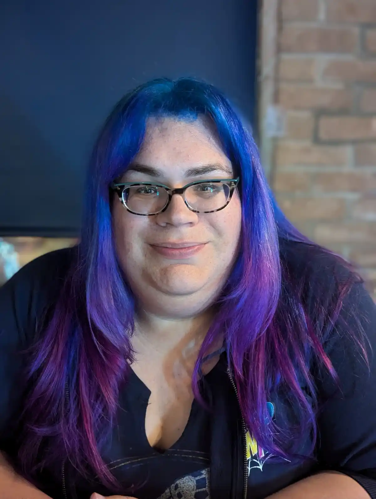 A person with long, vibrant blue and purple hair and glasses smiles while wearing a black zip-up jacket over a dark shirt…