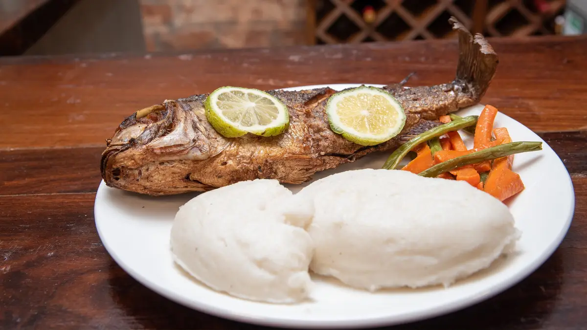 A whole fried fish on a white plate, with two lime slices placed over its eyes, resembling a face. The plate also contains…