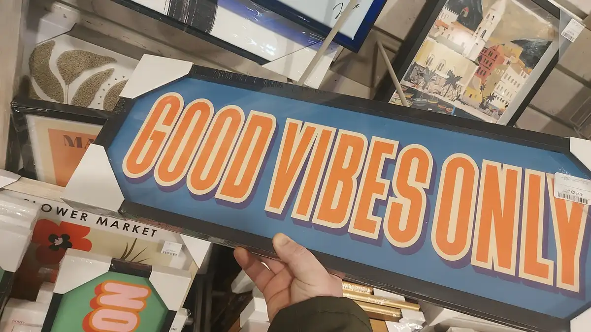 A hand holds a blue, arrow-shaped framed sign with bold orange text reading 'GOOD VIBES ONLY' in a store aisle surrounded by…