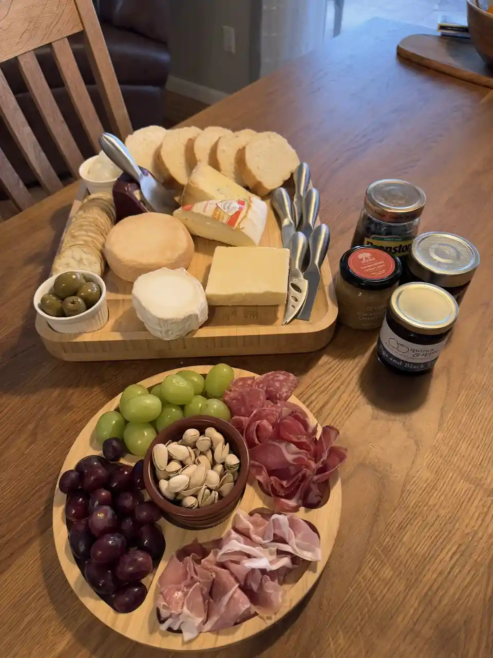 A wooden table displays two charcuterie boards: one rectangular with assorted cheeses, sliced bread, crackers, olives, and…