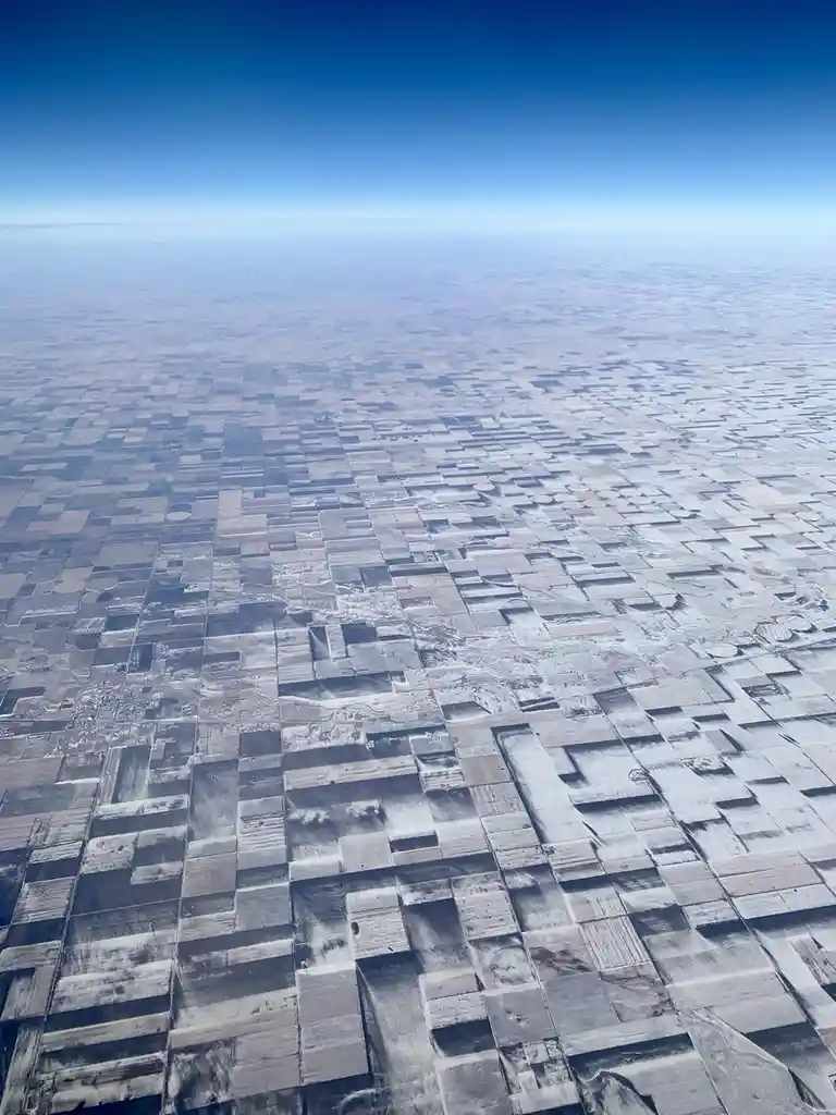 An aerial view from an airplane window shows a vast landscape of patchwork farmland, many fields covered in snow, under a…