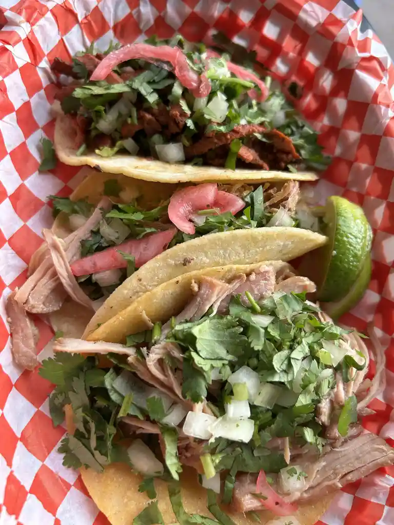 Three tacos filled with meat, cilantro, onions, and pickled onions sit in a basket lined with red and white checkered paper…