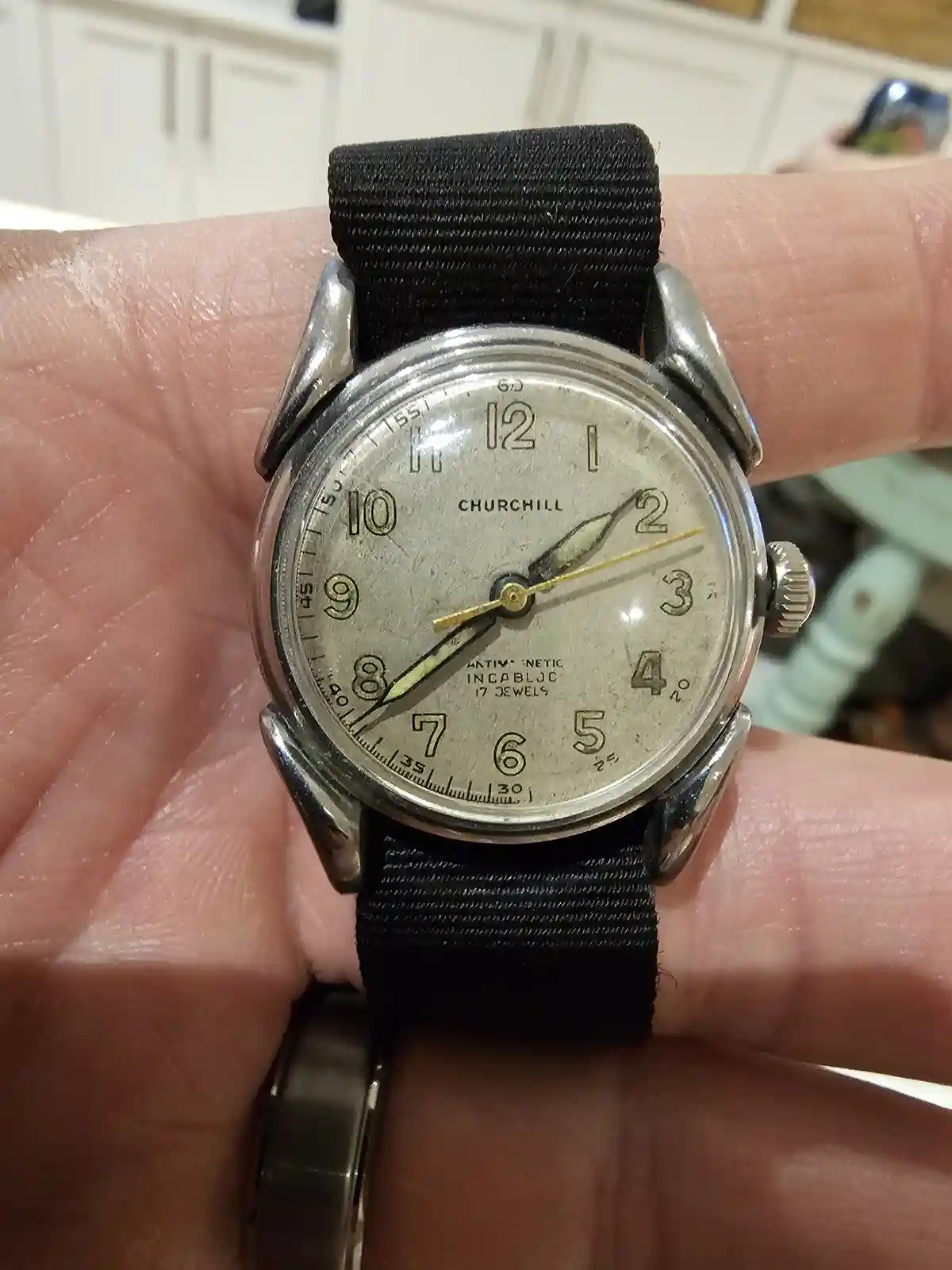 A close-up photo shows a person's hand holding an old, silver-toned Churchill wristwatch with a black fabric strap. The…