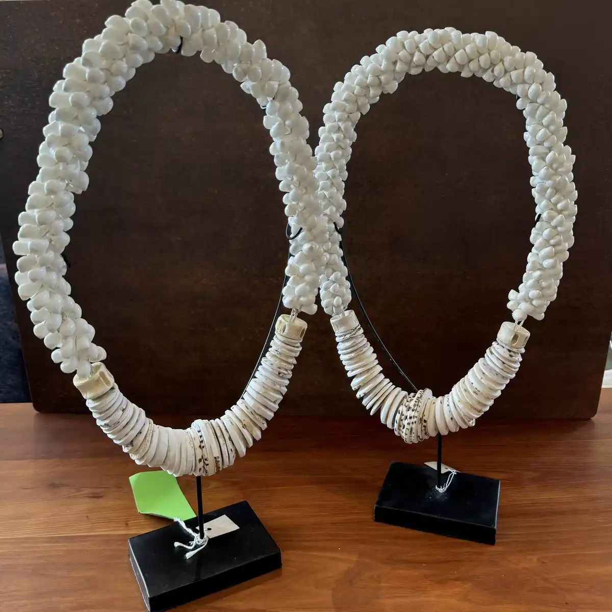Two ornate, white-beaded necklaces shaped like the letter 'O' are displayed on black stands on a wooden surface. The…