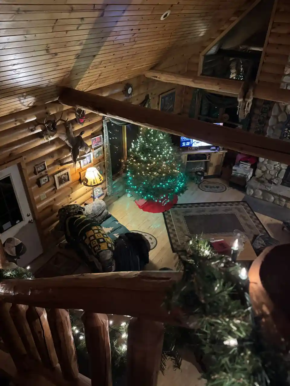 An overhead view of a cozy, wood-paneled log cabin living room decorated for Christmas, featuring a lit tree, rustic…