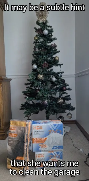 A decorated artificial Christmas tree stands in a corner, with several boxes of HDX brand trash bags stacked at its base…