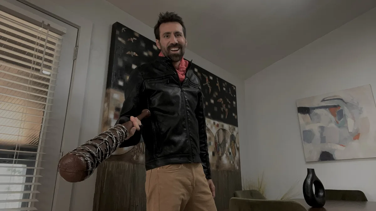 A man with a beard and black leather jacket stands in a modern living room holding a barbed-wire-wrapped baseball bat…