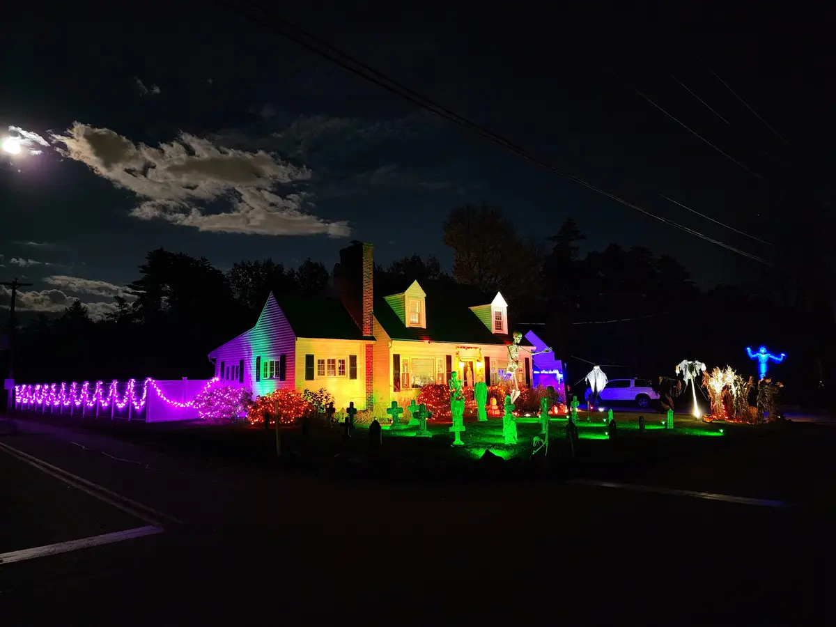 A house at night is extravagantly decorated with Halloween lights and props, including glowing skeletons, tombstones, and a…