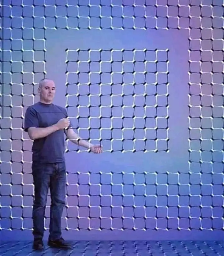 A man in a dark t-shirt and jeans stands in front of a large, glowing blue grid background that forms the shape of the…