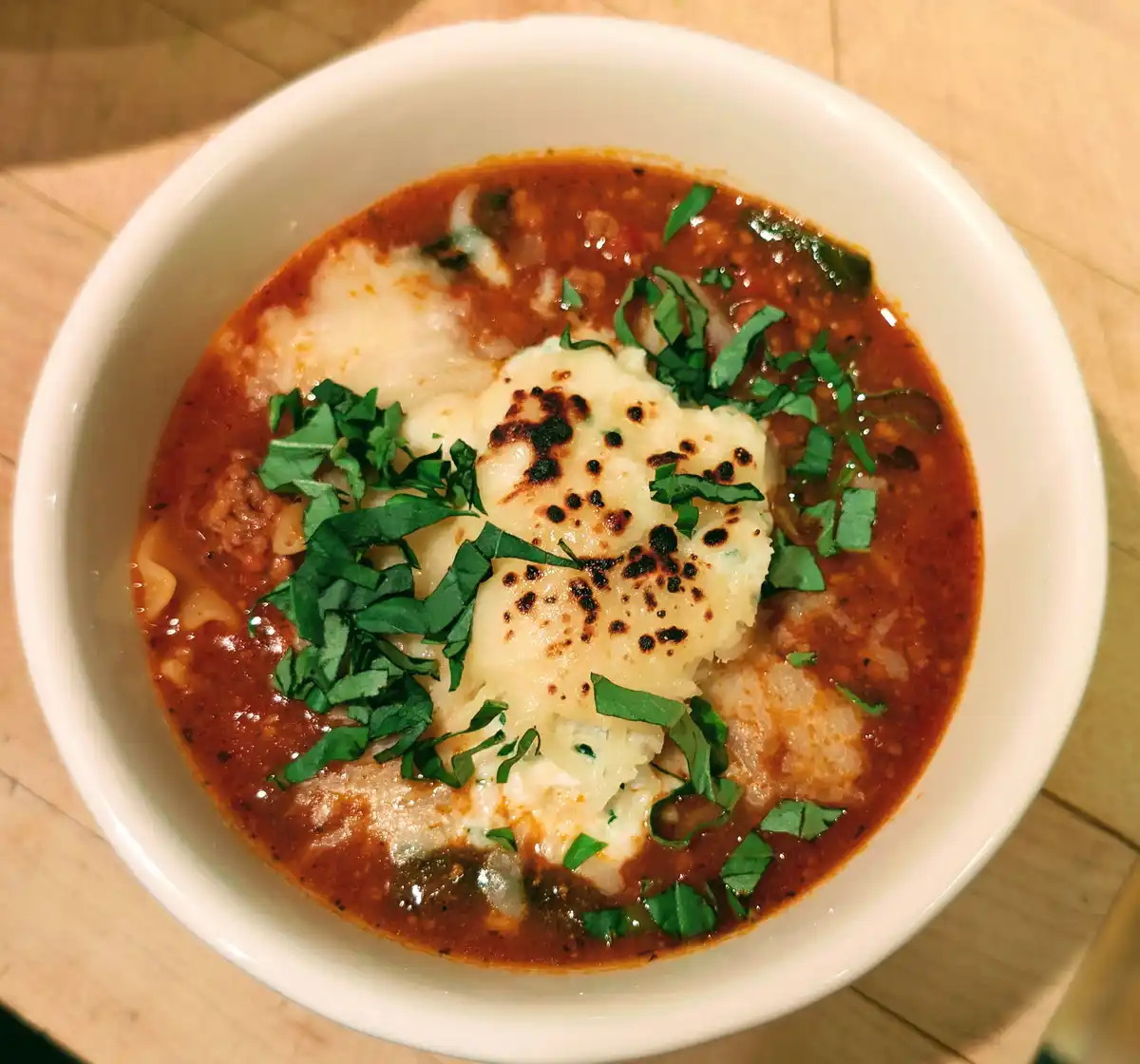A top-down photo of a white bowl filled with a red tomato-based soup or stew, topped with a large, toasted, creamy white…