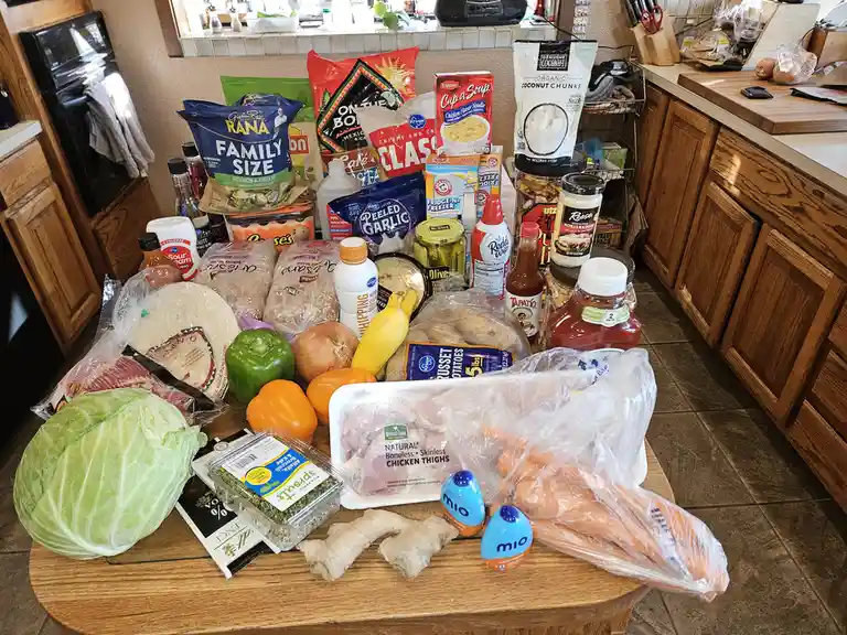 A kitchen counter is covered with a massive, chaotic pile of groceries including chicken thighs, carrots, cabbage, bell…