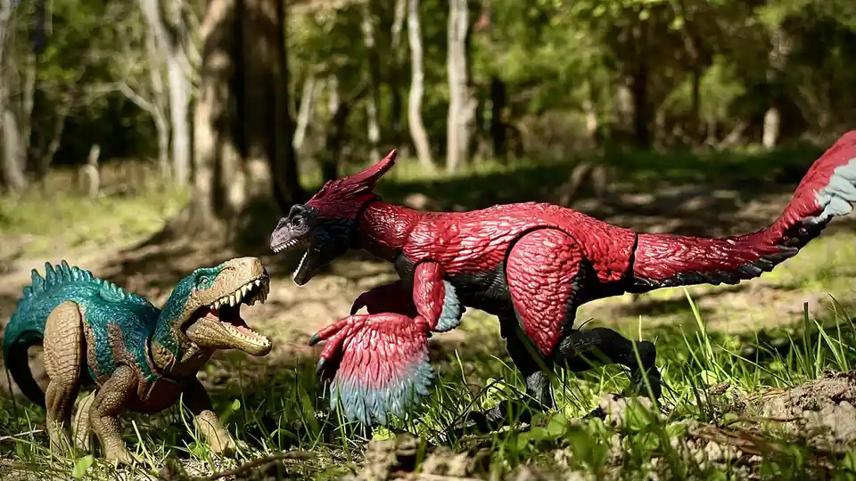 Two plastic dinosaur action figures, one teal and one red, are posed in a tense face-off in a sunlit forest setting. The red…