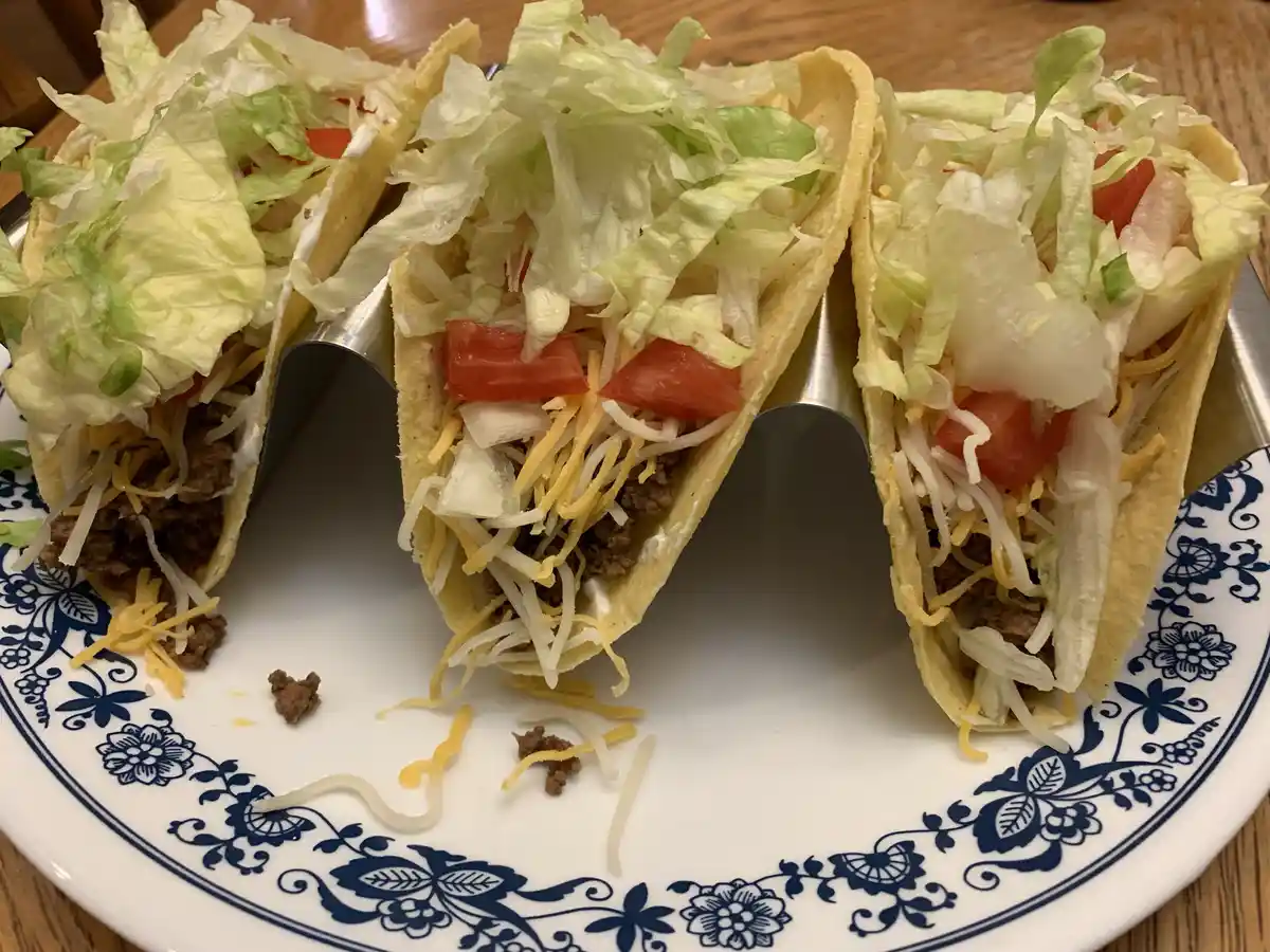 Three soft-shell tacos filled with ground beef, shredded cheese, diced tomatoes, onions, and shredded lettuce are arranged…