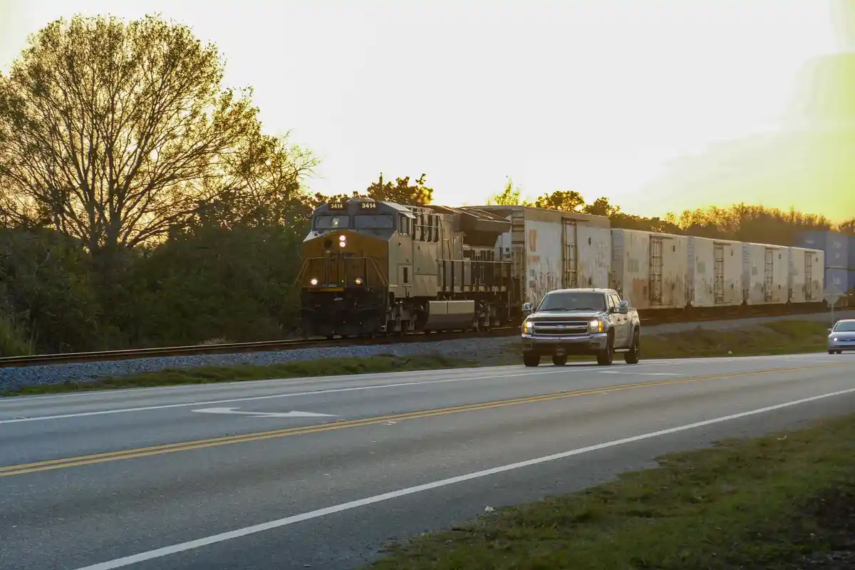 A silver pickup truck drives on a road parallel to a freight train at sunset. The train, with locomotive number 3414, is…