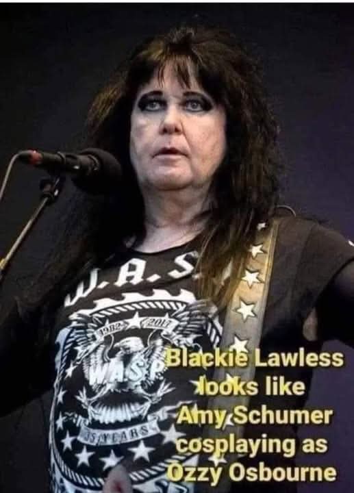 A woman with dark, shaggy hair and heavy eye makeup stands in front of a microphone wearing a black W.A.S.P. band t-shirt…