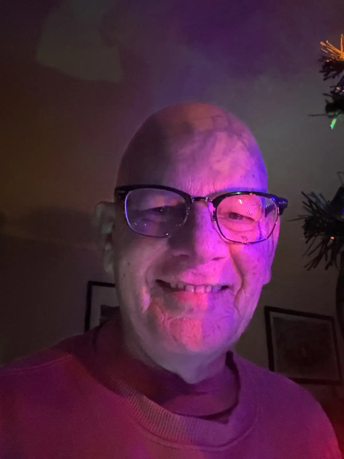 A close-up photo of a bald man with glasses smiling, bathed in purple and pink lighting, with a Christmas tree visible in…