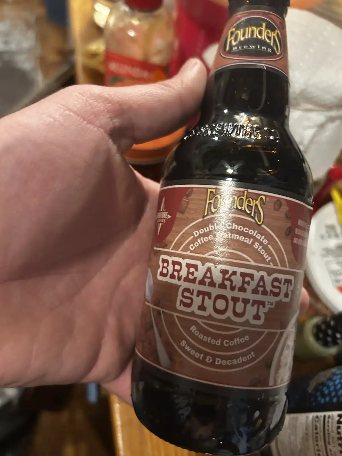 A hand holds a dark glass bottle of Founders Brewing's 'Breakfast Stout' beer, which is labeled as 'Double Chocolate Coffee…