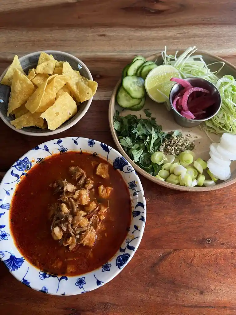 A top-down photo shows a bowl of red, chunky stew with shredded meat, accompanied by a plate of garnishes including sliced…