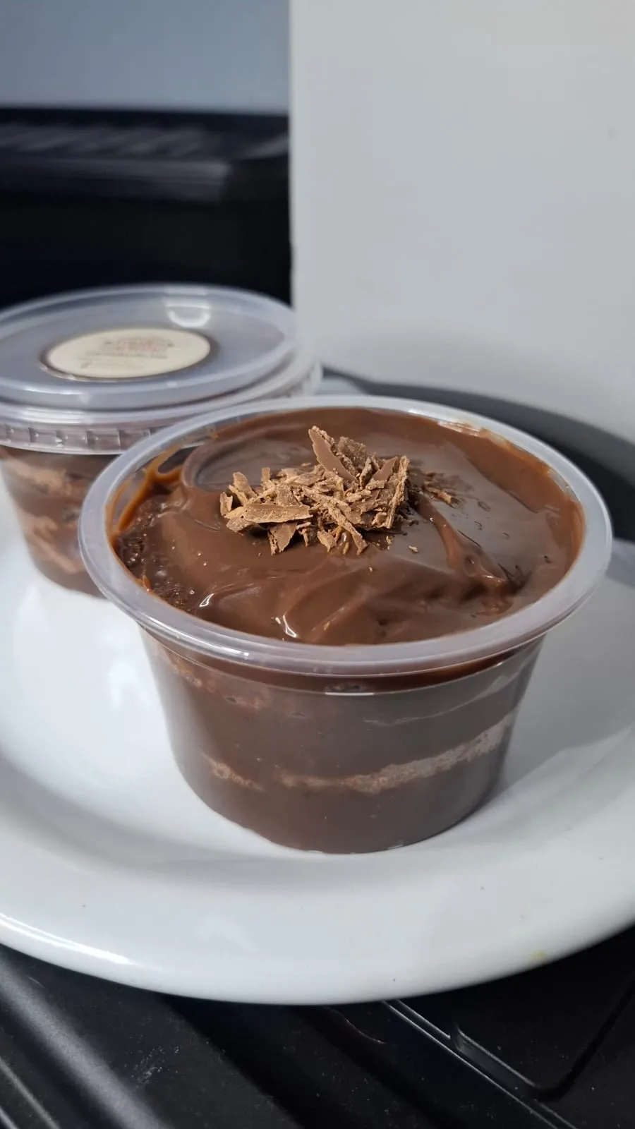 A close-up photo shows two plastic cups of chocolate mousse dessert on a white plate, with the front cup topped with…