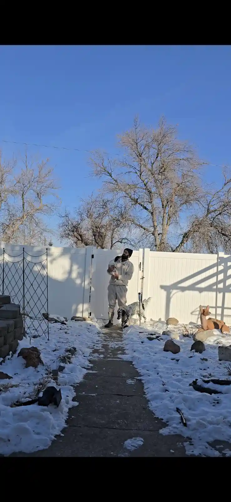 A man in a light-colored sweatsuit and beanie stands in a snowy backyard, holding a small white dog while a larger spotted…