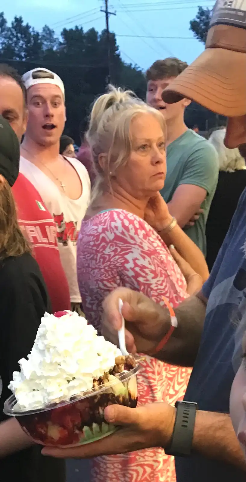 A woman with blonde hair in a ponytail, wearing a pink and white patterned top, looks unimpressed while standing in a crowd…