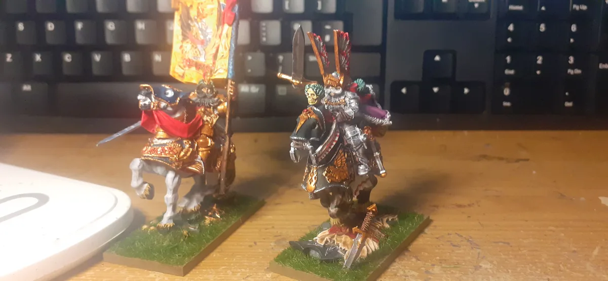 Two intricately painted fantasy miniatures of armored knights on horseback sit on grassy bases on a wooden desk, with a…
