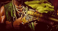A man in a green vest and multiple necklaces plays a saxophone, transitioning from a close-up of his hands on the instrument…