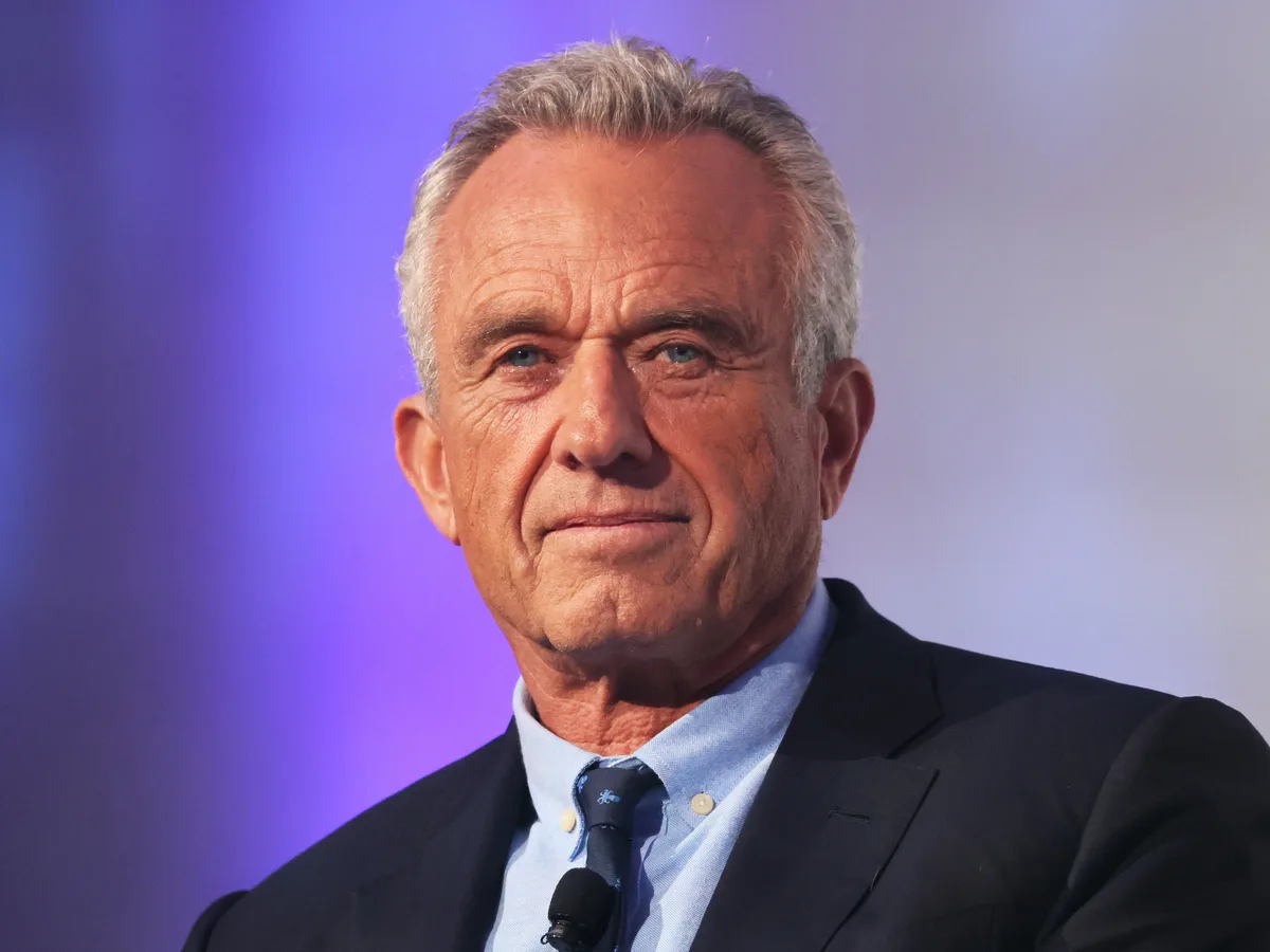 A close-up photo of a man with gray hair, wearing a dark suit and light blue shirt, looking directly at the camera with a…